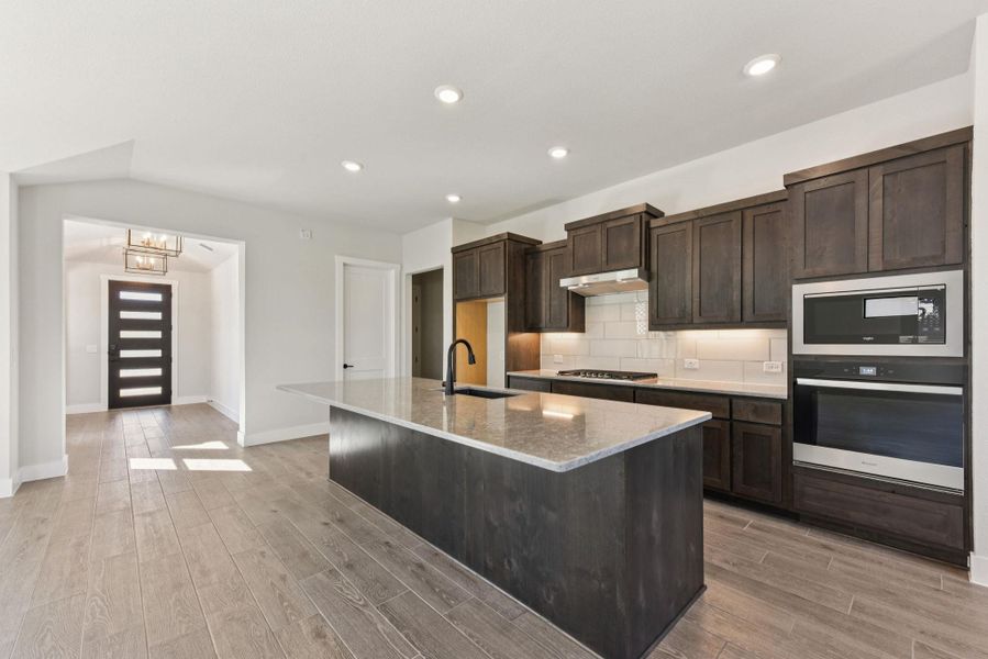 Kitchen with backsplash, appliances with stainless steel finishes, dark brown cabinetry, a chandelier, and light stone countertops Kitchen with backsplash, appliances with stainless steel finishes, dark brown cabinetry, a chandelier, and light stone countertops