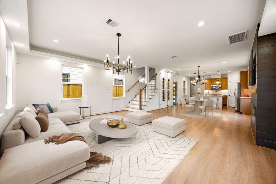 Spacious open-concept living area featuring warm wood flooring, recessed lighting, and abundant natural light. The focal point is a sleek linear fireplace with a modern accent wall and floating mantle. A tray ceiling with contemporary chandelier adds architectural interest, while the seamless flow into the dining and kitchen areas makes this space ideal for everyday living and entertaining.