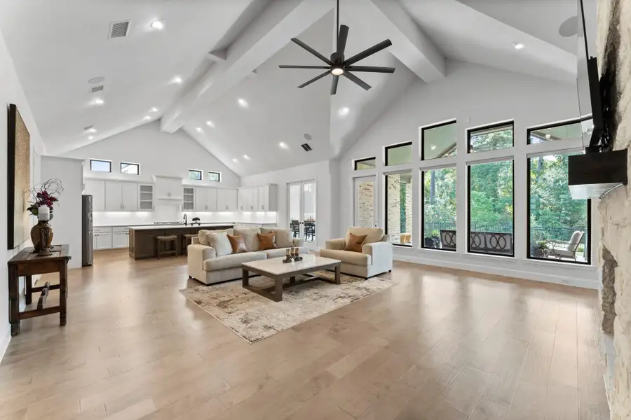 A wide shot of the open-concept living area. The view extends from the fireplace on the right to the kitchen on the left. The light wood floors connect the two spaces seamlessly. The room features high, angled ceilings with exposed beams and a large ceiling fan. A wide shot of the open-concept living area. The view extends from the fireplace on the right to the kitchen on the left. The light wood floors connect the two spaces seamlessly. The room features high, angled ceilings with exposed beams and a large ceiling fan.