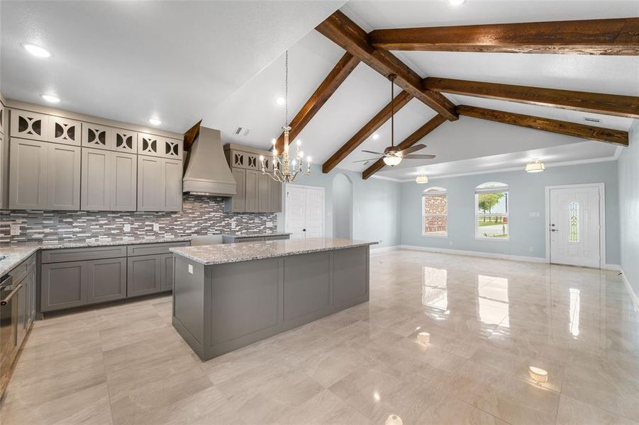 Kitchen with beamed ceiling, light stone counters, decorative light fixtures, ceiling fan with notable chandelier, and custom exhaust hood