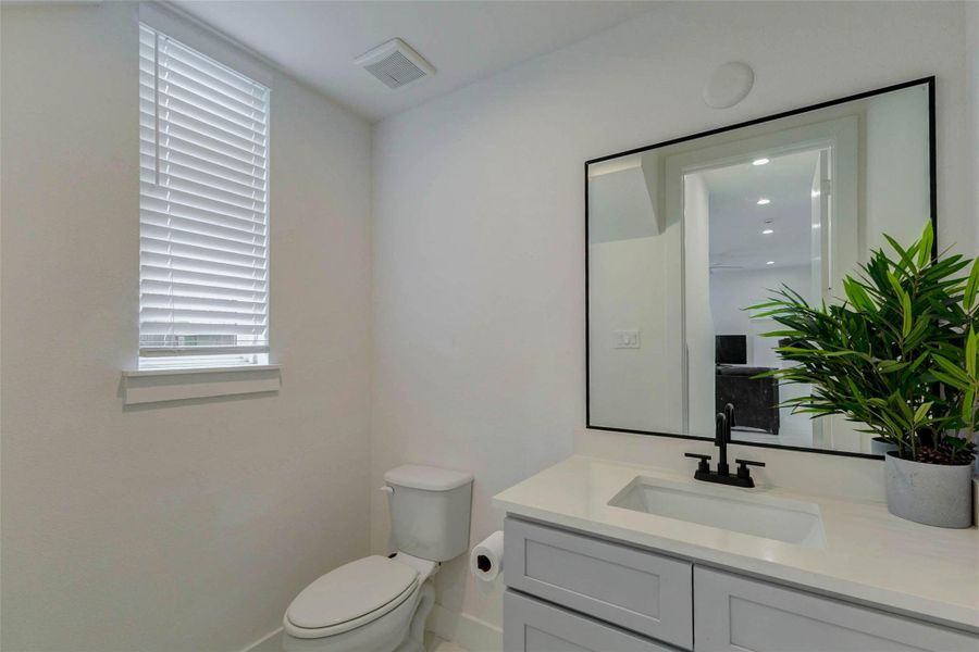 Stylish powder bath with a clean modern vanity, light countertop, undermount sink, black faucet, framed mirror, neutral walls, window with blinds, and simple contemporary finishes. A fresh, bright guest bath that continues the home’s modern white-and-black design palette.