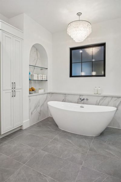 Another view of the 71" Soaking Tub with a Wall-Mounted Tub Filler in Polished Chrome and a Tiered Chandelier with Natural White Shell. There is also open glass shelving in the alcove wall that holds neatly stacked towels, jars, and bath accessories. Linen Storage Closet for Toiletries and Towels.