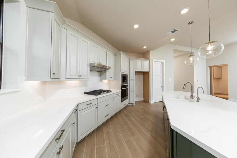 The open-concept gourmet kitchen designed for both cooking and entertaining. It features elegant white cabinetry, pristine quartz countertops, and a large central island that doubles as a functional breakfast bar. Three glass pendants elegantly hang above the kitchen island, providing focused task lighting and a touch of refined style.