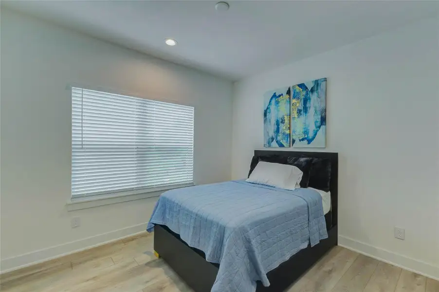 Bright secondary bedroom with light wood-look flooring, large window with blinds, recessed lighting, neutral walls, and a comfortable room layout. The space offers a clean, modern feel with flexibility for a bedroom, guest space, office, or other everyday use.