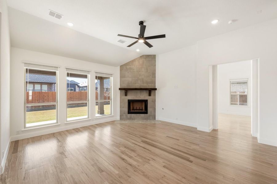 Open living room with tile fireplace, hardwood floors, ceiling fan, and large windows overlooking backyard