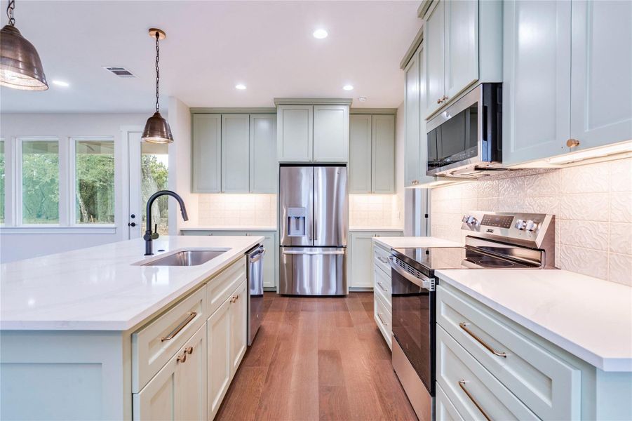 Kitchen with stainless steel appliances, pendant lighting, light stone countertops, a kitchen island with sink, and light wood-style flooring