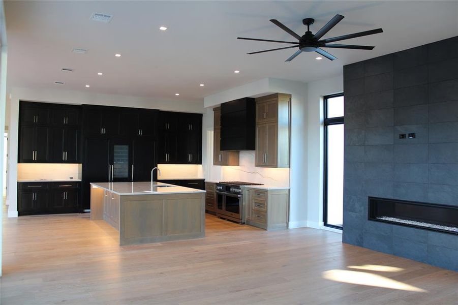 Kitchen featuring light wood-type flooring, ventilation hood, a tile fireplace, a center island with sink, and stainless steel stove