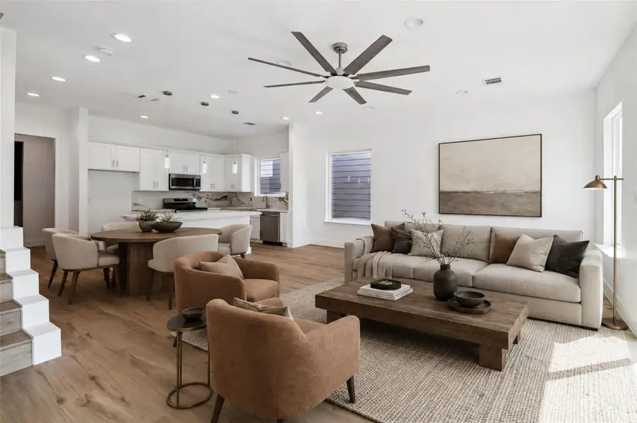 From this angle, the open layout between the living area and kitchen truly shines. A spacious footprint allows plenty of room for comfortable seating and dining arrangements, while the large ceiling fan adds style and everyday comfort. Natural light pours in through expansive windows and glass doors, highlighting the warm flooring and crisp neutral walls. The kitchen remains visually connected for easy conversation and entertaining. This photo has been virtually staged for visual reference.