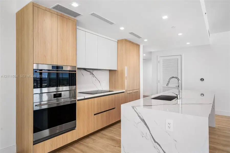 Kitchen featuring wood-finish cabinetry, white slab-style upper cabinets, stone-surfaced island with waterfall edge, integrated stainless steel oven and microwave, and recessed ceiling lighting Kitchen featuring wood-finish cabinetry, white slab-style upper cabinets, stone-surfaced island with waterfall edge, integrated stainless steel oven and microwave, and recessed ceiling lighting