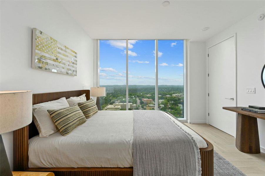 Serene secondary bedroom with mesmerizing sunset views. Floor-to-ceiling windows frame a spectacular panorama of the city and surrounding landscape, creating a captivating backdrop for peaceful evenings. The tranquil space features warm wood accents, soft neutral tones, and thoughtful lighting that complements the natural glow of golden hour, offering the perfect retreat after a busy day.