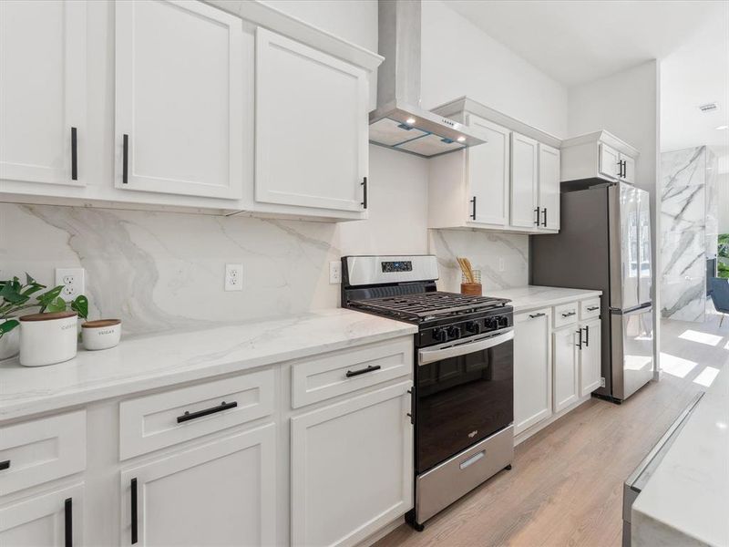 Kitchen with light stone counters, stainless steel appliances, white cabinets, light wood-type flooring, and wall chimney exhaust hood