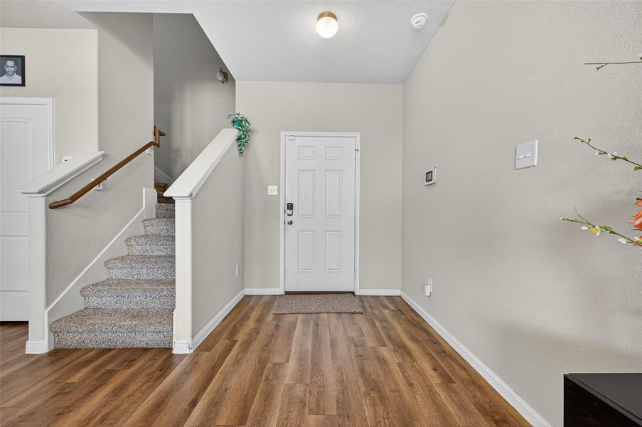 This photo showcases a bright entryway with wood flooring, neutral walls, and a carpeted staircase leading to the upper level. The space is well-lit and features a clean, modern design.