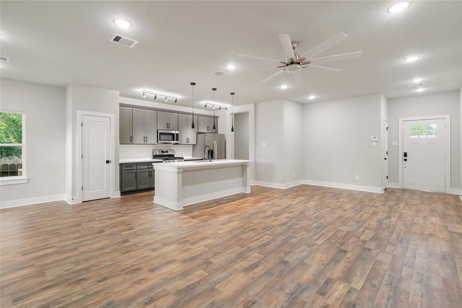 This photo showcases a modern, open-concept living area with wood-style flooring and recessed lighting. The kitchen features stainless steel appliances, pendant lights, and a large island with seating. Neutral walls and a ceiling fan add to the spacious, inviting atmosphere. This photo showcases a modern, open-concept living area with wood-style flooring and recessed lighting. The kitchen features stainless steel appliances, pendant lights, and a large island with seating. Neutral walls and a ceiling fan add to the spacious, inviting atmosphere.