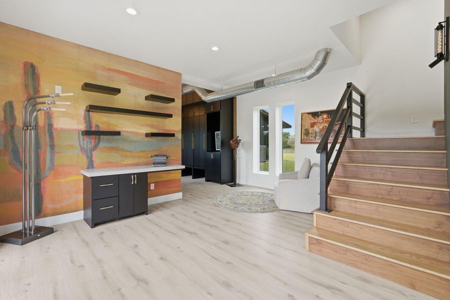 This modern space features a bright room with light wood flooring and an artistic accent wall. It includes sleek shelving, a small desk area, and a contemporary staircase. Large windows provide natural light, and a cozy seating area adds comfort.