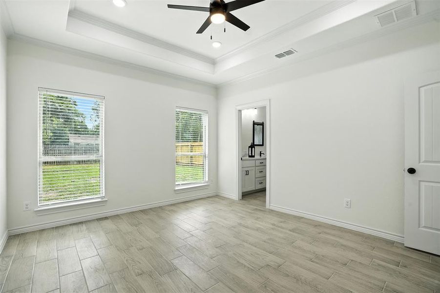 Unfurnished bedroom featuring a raised ceiling, wood finish floors, ensuite bathroom, crown molding, and ceiling fan