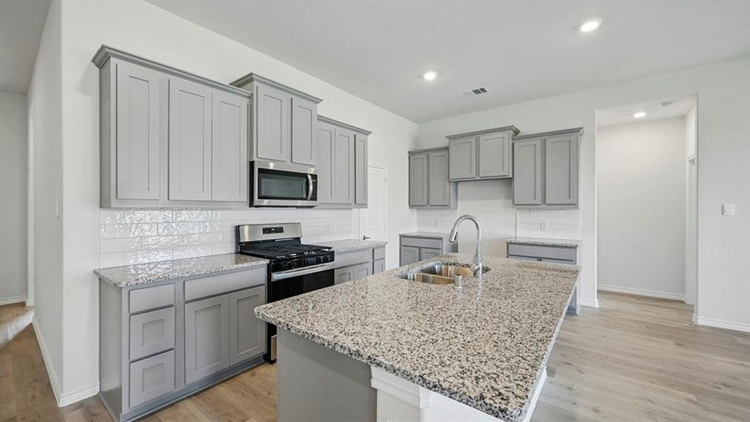Kitchen featuring gray cabinets, decorative backsplash, light stone countertops, a center island with sink, and recessed lighting