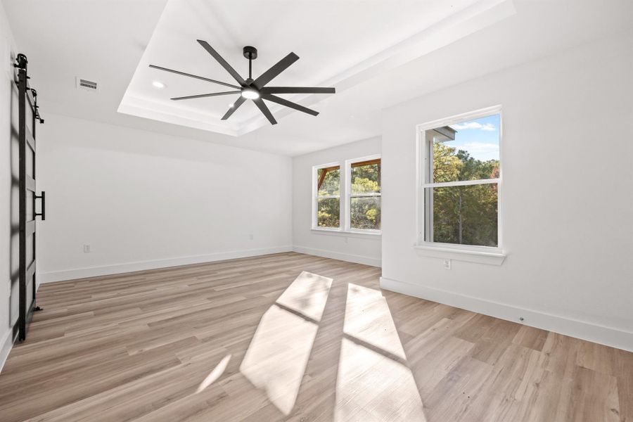 Primary bedroom with a barn door leading to the primary bath, light wood-style flooring, ceiling fan, and a lighted tray ceiling