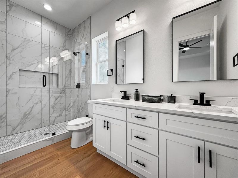Full bathroom with a marble finish shower, double vanity, toilet, wood finished floors, and ceiling fan