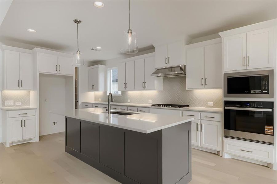 Kitchen with stainless steel appliances, two tone color scheme, an island with sink, decorative light fixtures, and light wood-style flooring