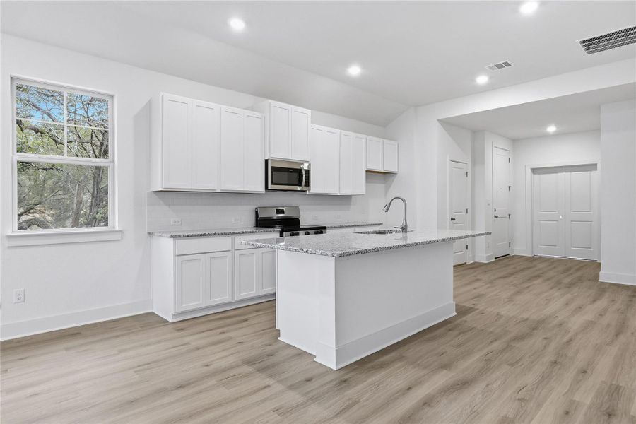 Kitchen with stainless steel appliances, light stone countertops, white cabinetry, a center island with sink, and light wood-style flooring