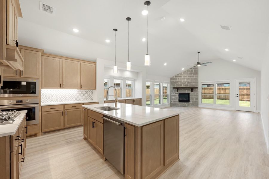 Open kitchen with wood cabinets, large island, and vaulted ceiling open to living room with brick fireplace