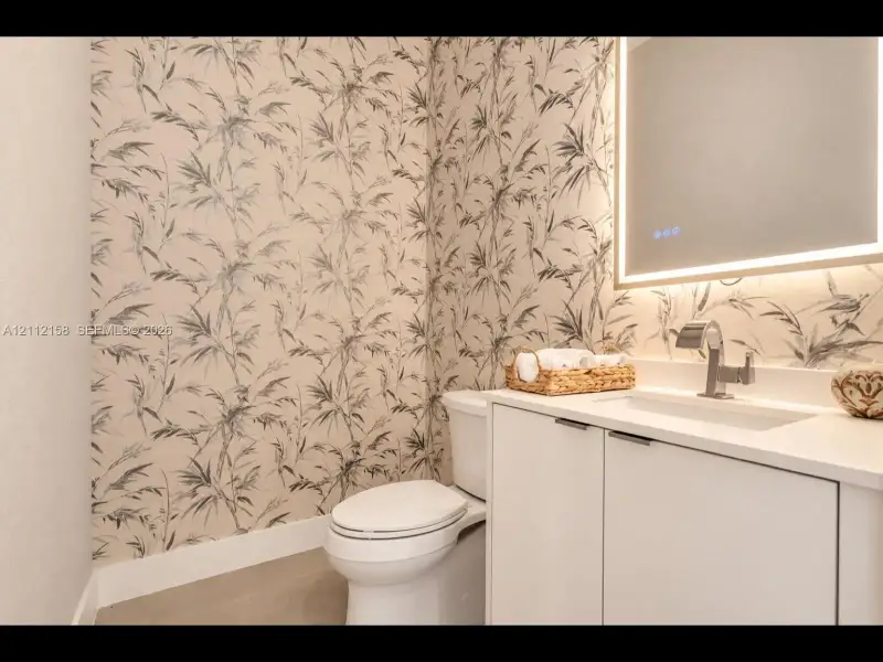 Stylish bathroom with designer patterned wallpaper, white vanity, chrome fixtures, and a modern LED-backlit mirror