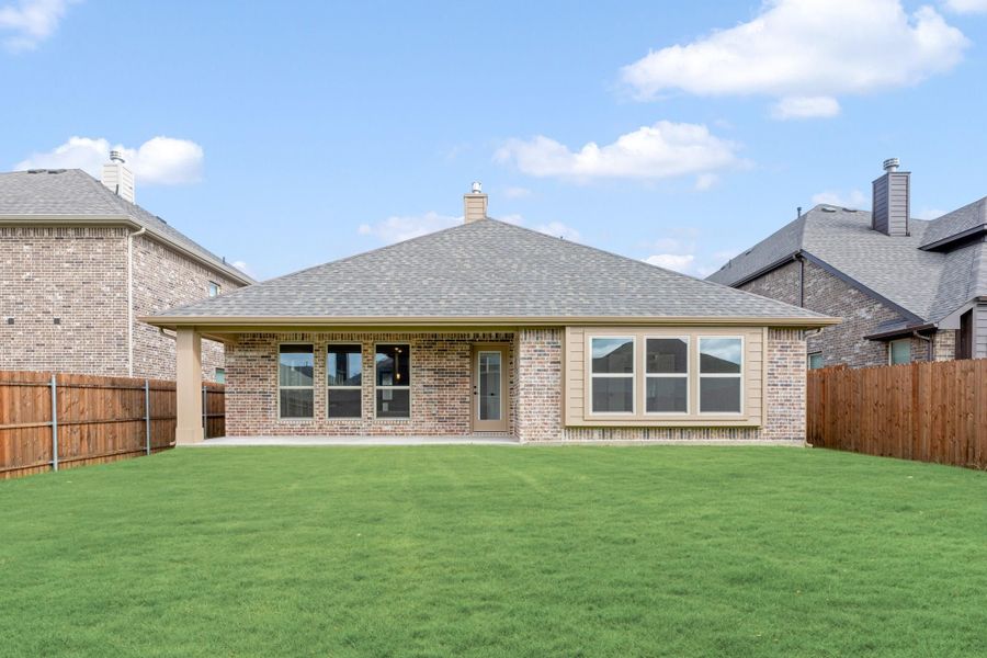 Rear view of single-story brick home with covered patio, large windows, and fenced green backyard
