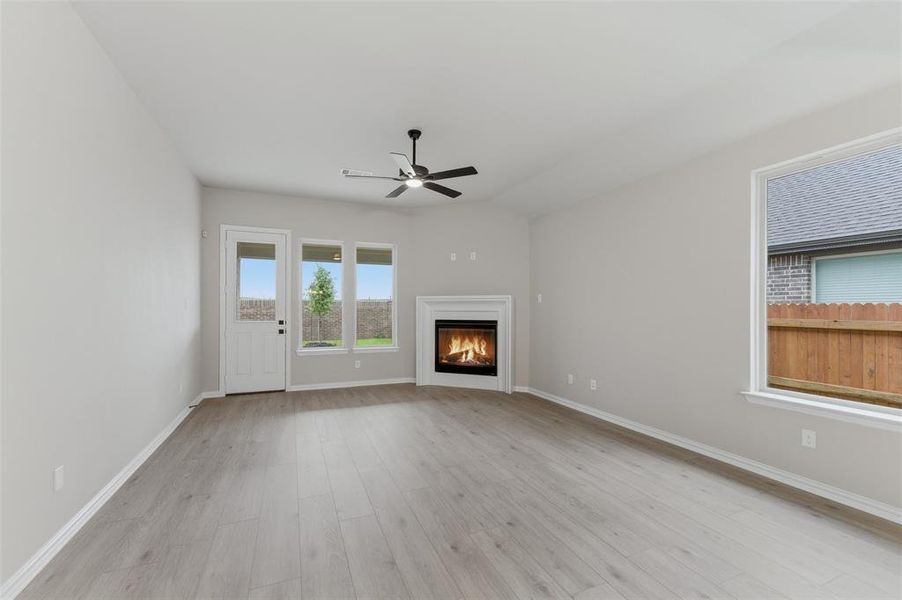Spacious interior featuring a corner fireplace with white mantel, a ceiling fan with integrated lighting, wood-finish flooring, and multiple windows providing natural light