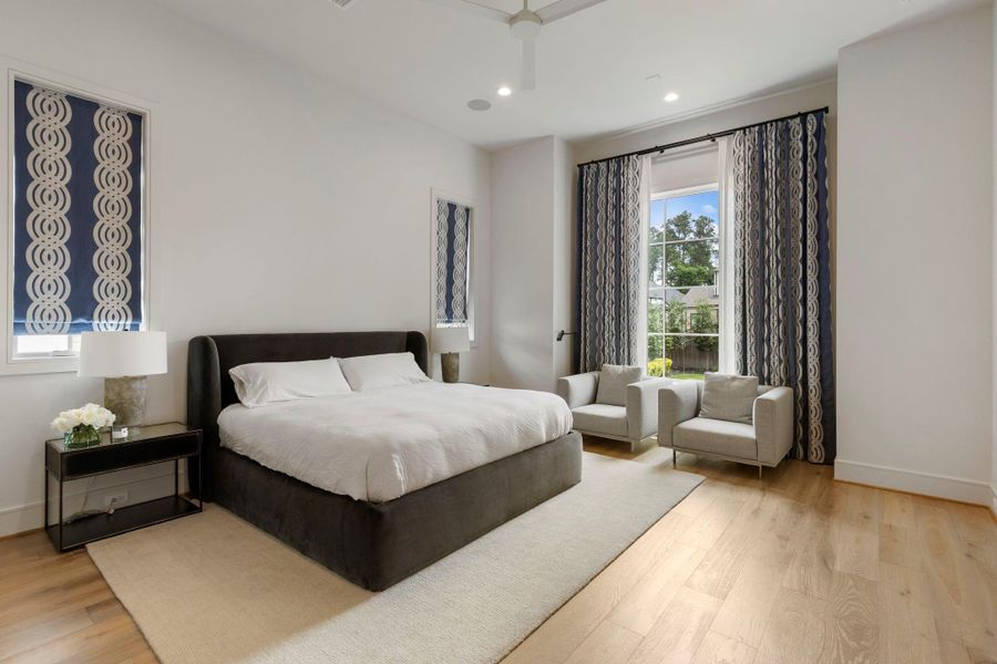 Beautiful primary bedroom retreat with wide plank hardwood flooring, designer drapery, and a cozy sitting area positioned beside large windows that bring in natural light and peaceful backyard views.