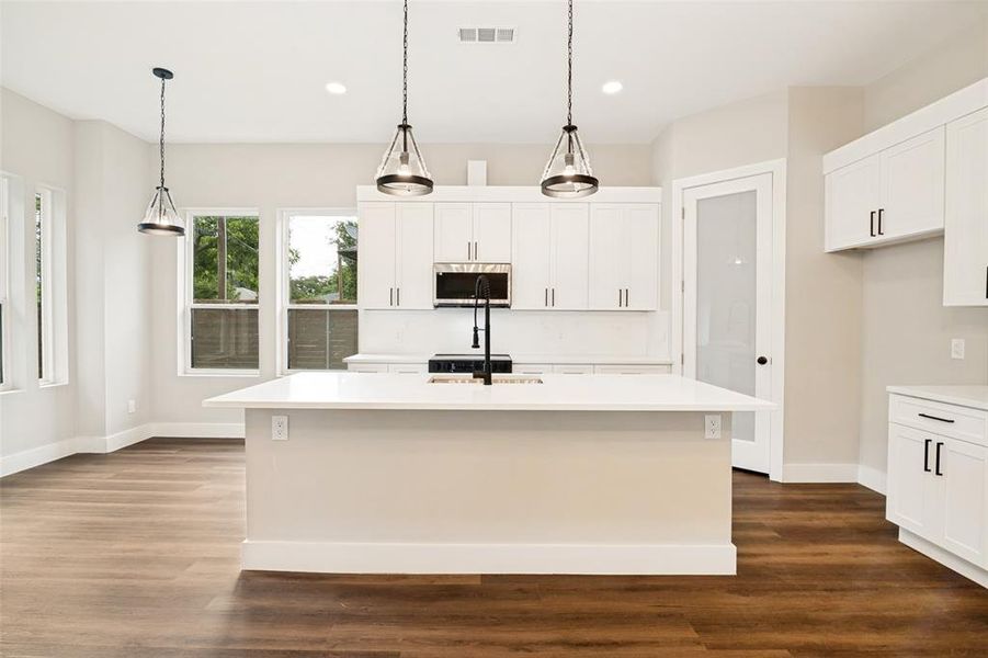 Contemporary kitchen featuring an expansive central island with a solid surface countertop, white shaker cabinetry with black hardware, wood-finish flooring, and recessed and pendant lighting