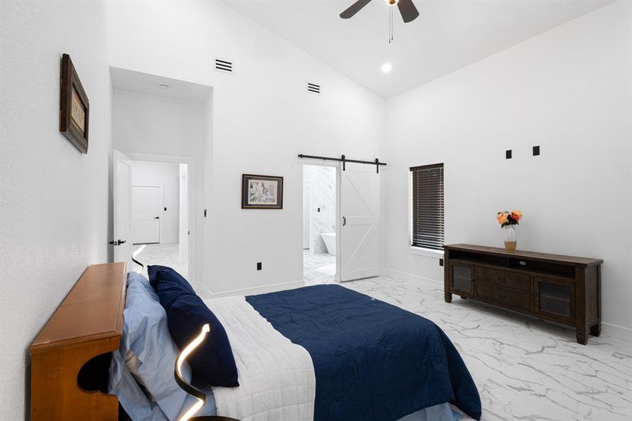 Bedroom featuring a barn door, light marble finish flooring, high vaulted ceiling, ceiling fan, and ensuite bath