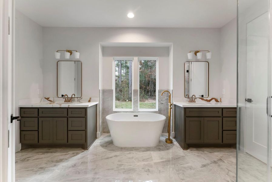 This primary en-suite features a modern, elegant design with a freestanding tub centered between dual vanities. Natural light flows in through a large window, highlighting the sleek fixtures and sleek  flooring.
