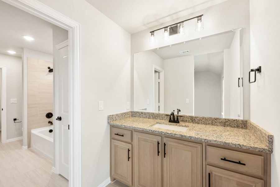 Bathroom with wood vanity, granite countertop, large mirror, and soaking tub visible through doorway Bathroom with wood vanity, granite countertop, large mirror, and soaking tub visible through doorway