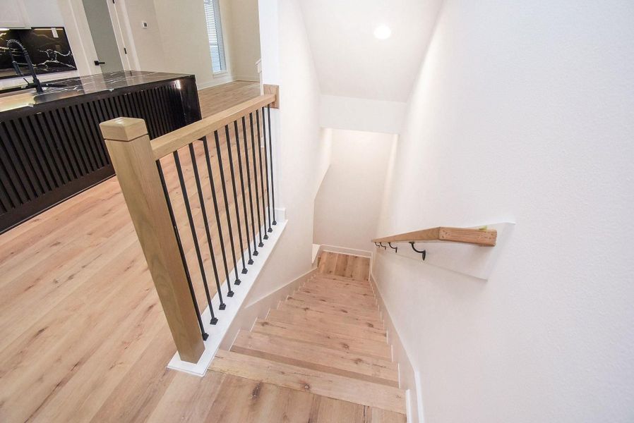 This photo shows a modern staircase with light wood floors and a sleek black railing. The area is well-lit, leading down from an open-concept space featuring a stylish kitchen island.