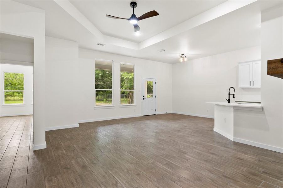 Spacious living area featuring wood-finish flooring, a tray ceiling with a modern ceiling fan, and multiple windows providing natural light