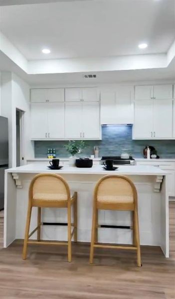 Kitchen featuring a white island with corbel details, wood-finish flooring, white cabinetry, a light blue tile backsplash, and recessed lighting