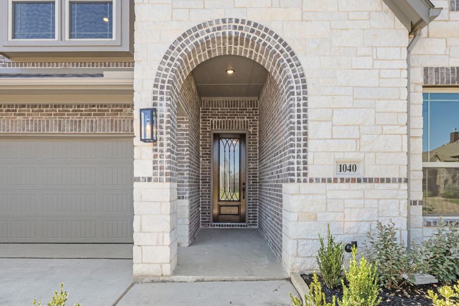 Home entrance with arched brick alcove, iron-glass front door, stone exterior, and wall lantern beside garage Home entrance with arched brick alcove, iron-glass front door, stone exterior, and wall lantern beside garage