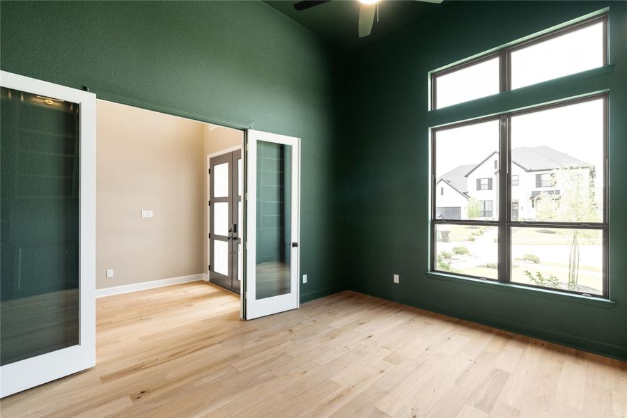 A spacious study with elegant green walls features large windows for abundant natural light and sleek glass double doors, complemented by light wood flooring for a modern and inviting atmosphere.