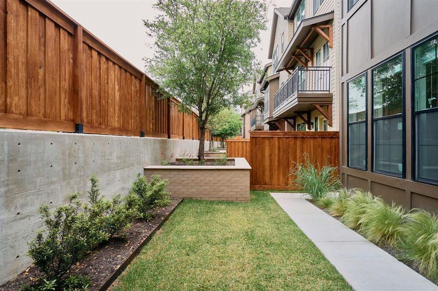 Private outdoor space featuring a concrete retaining wall, wood fencing, a grass lawn, and a paved walkway Private outdoor space featuring a concrete retaining wall, wood fencing, a grass lawn, and a paved walkway