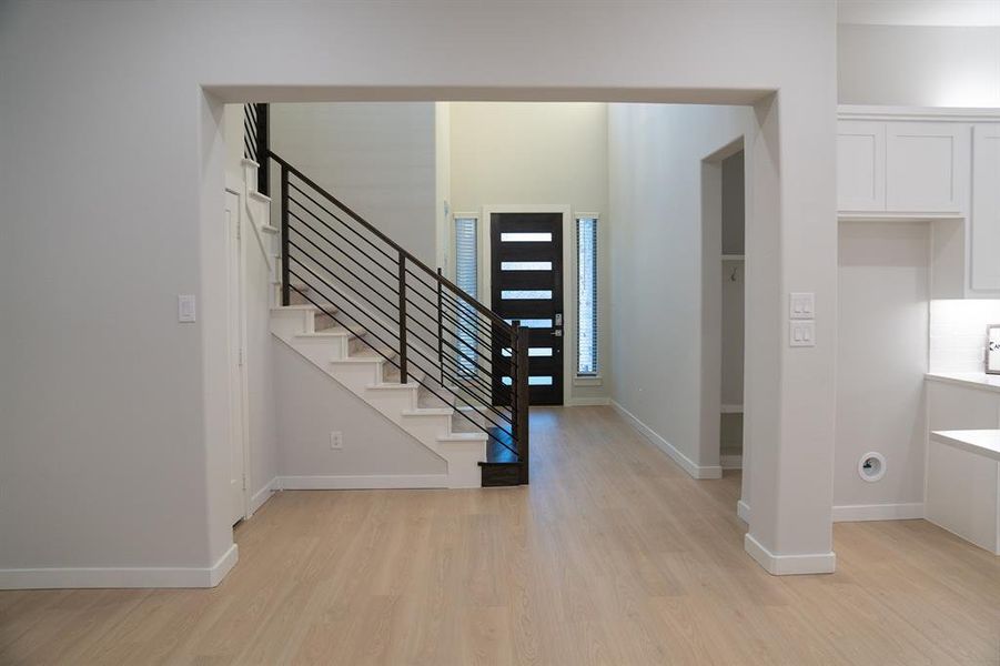 Open concept living space featuring light wood-finish flooring, a modern black entry door with frosted glass panels, and a sleek staircase with black metal balusters Open concept living space featuring light wood-finish flooring, a modern black entry door with frosted glass panels, and a sleek staircase with black metal balusters