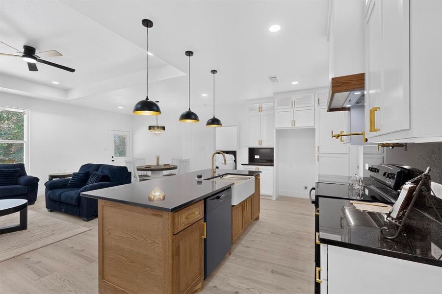 Open-concept kitchen featuring an oversized island with a farmhouse sink, black granite-style countertops, white cabinetry with gold-tone hardware, and light wood-finish flooring