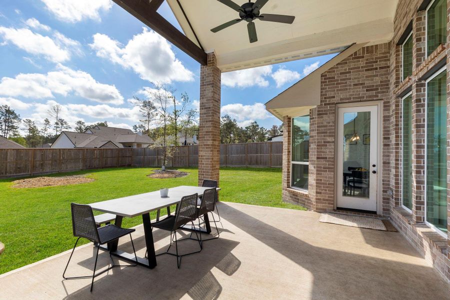 This photo features a spacious, covered patio with a ceiling fan, perfect for outdoor dining and entertaining. It overlooks a large, well-maintained backyard with a tall privacy fence, providing a serene and private setting. The patio is connected to the house with large windows and a door, enhancing indoor-outdoor flow.