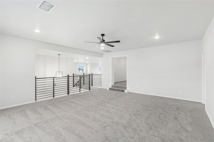 Open upstairs loft with gray carpet, ceiling fan, and metal stair railing overlooking lower level
