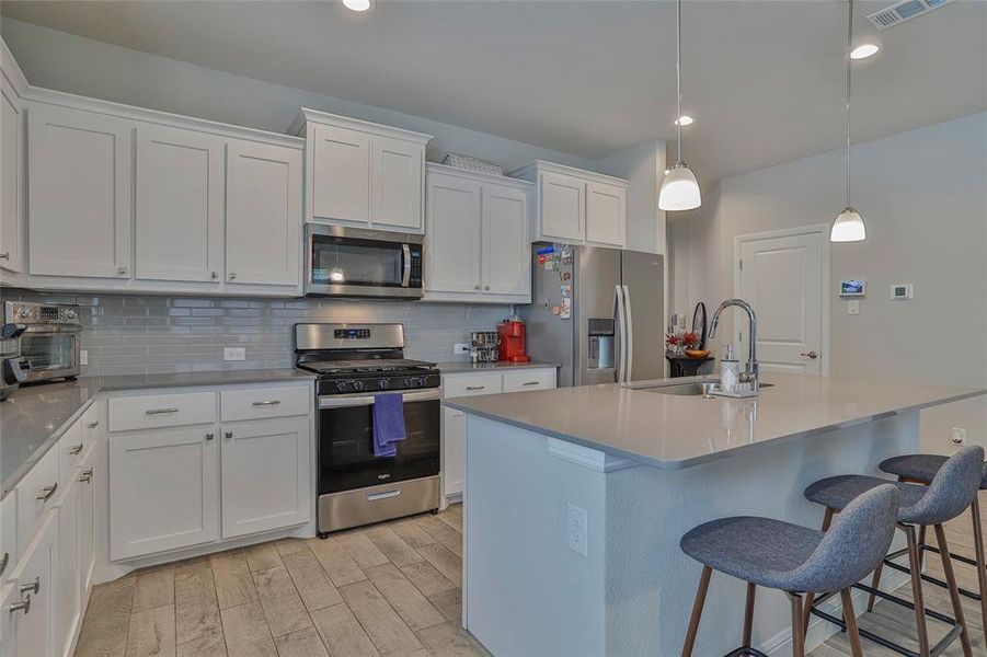 Kitchen with stainless steel appliances, decorative backsplash, white cabinetry, a kitchen island with sink, and light wood-type flooring Kitchen with stainless steel appliances, decorative backsplash, white cabinetry, a kitchen island with sink, and light wood-type flooring