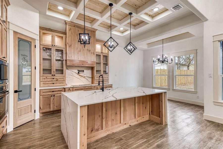 Kitchen with coffered ceiling, light stone countertops, a chandelier, wood tiled floors, and a kitchen island with sink