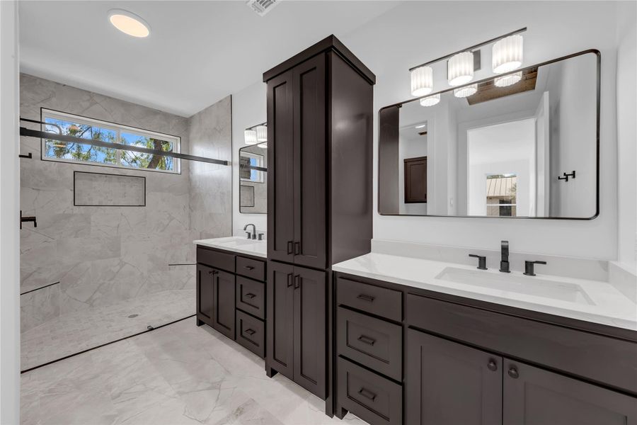 A contemporary primary bath showcases dark cabinetry, double vanities, and a walk-in shower with stone tile.