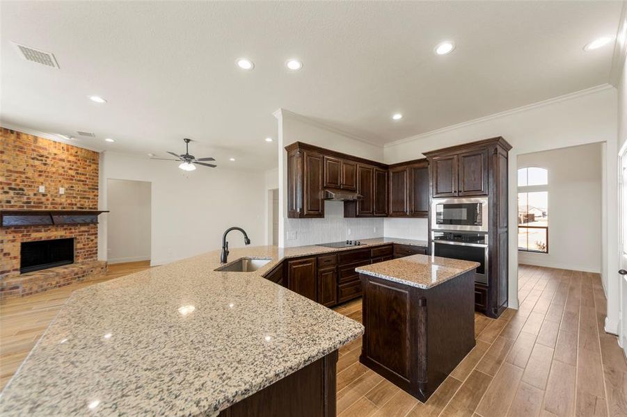 Kitchen featuring light wood-type flooring, stainless steel appliances, a brick fireplace, light stone countertops, and a kitchen island Kitchen featuring light wood-type flooring, stainless steel appliances, a brick fireplace, light stone countertops, and a kitchen island