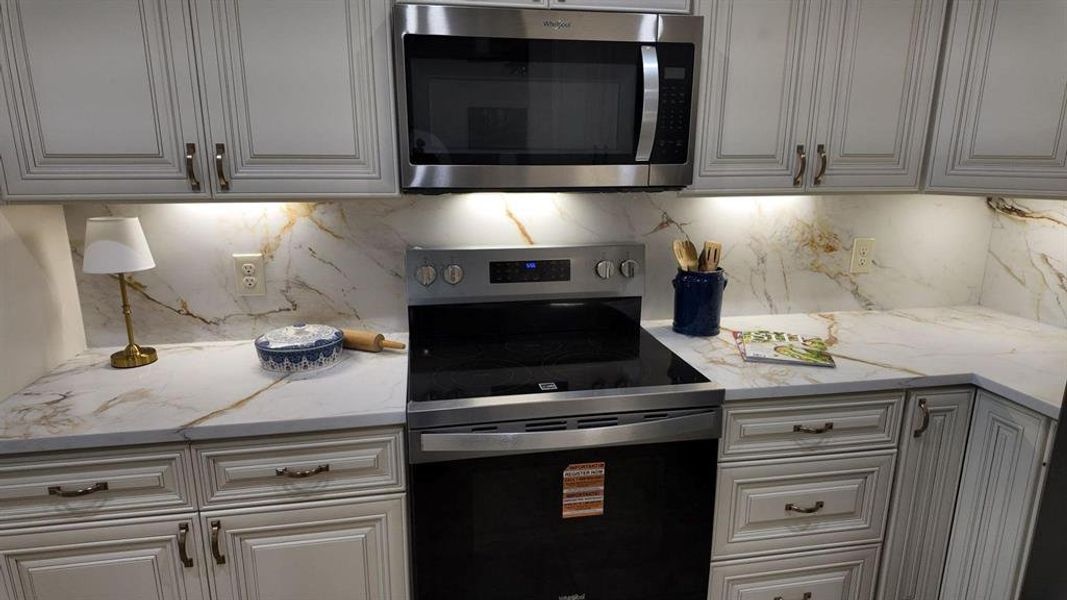 Kitchen featuring white shaker-style cabinetry, a white countertop with gold veining, a matching full-height backsplash, stainless steel appliances, and under-cabinet lighting
