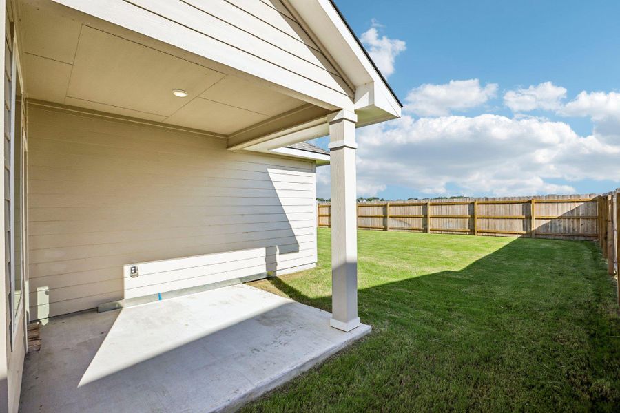A covered concrete patio overlooks a spacious grassy yard enclosed by a wooden privacy fence.