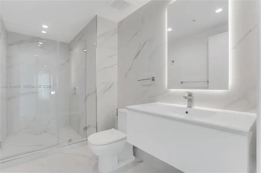 Modern bathroom featuring floor-to-ceiling large-format stone-patterned tile, a glass-enclosed walk-in shower, a white floating vanity with a seamless countertop, and a wall-mounted mirror with integrated backlighting